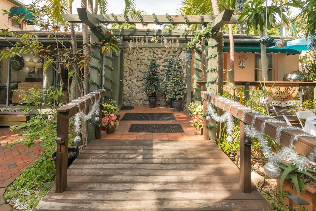 A wooden bridge leads through a lush garden, framed by greenery and festive decorations. A stone wall is visible at the end of the path, flanked by potted plants. Light filters softly through the nearby trees, creating a serene and inviting entryway.