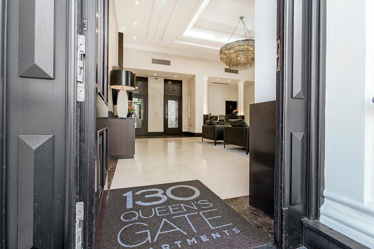 The entrance to 130 Queen's Gate Apartments features elegant design elements, such as a chandelier above and a welcoming reception area. A plush seating arrangement can be seen in the background, while the logo mat is visible at the doorway, inviting guests into the space.