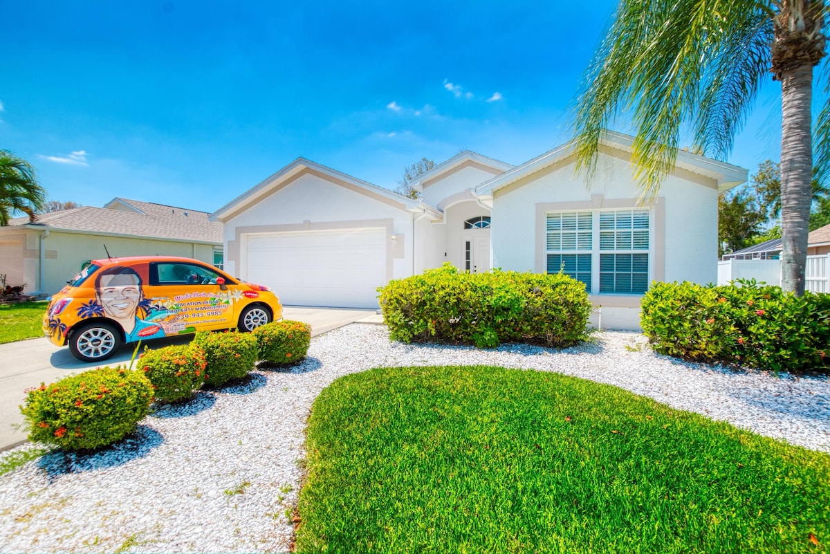 The exterior of the vacation home features a well-maintained lawn with lush green grass and neatly trimmed shrubs. A vibrant small car is parked in the driveway, adding a pop of color to the serene setting. The house showcases a spacious façade with large windows and a welcoming entrance.