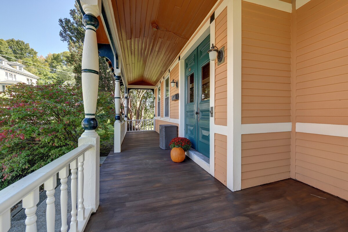 An inviting front porch features a wooden floor and is bordered by white railings. Two blue doors lead into the home. A decorative pumpkin and a pot of blooming flowers add a touch of seasonal charm to the space.