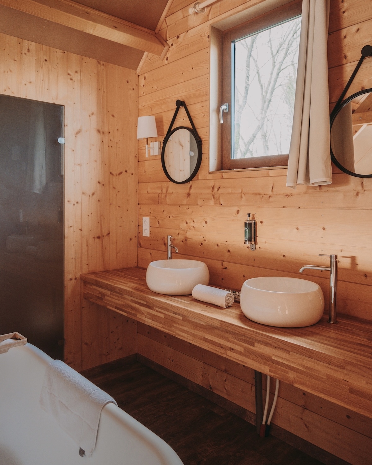 A wooden bathroom features a dual vanity with two round sinks placed atop a long, wide countertop. Natural light enters through the window, illuminating the wooden walls. A shower area with a glass partition is visible, along with two round mirrors above the sinks.
