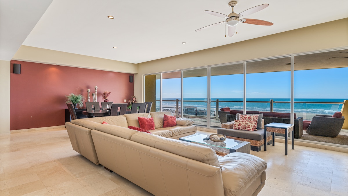A spacious living area features a large sectional sofa, complemented by decorative cushions. Large windows offer expansive views of the ocean, bathing the room in natural light. A dining table accommodates several guests, while the ceiling fan provides a comfortable atmosphere.