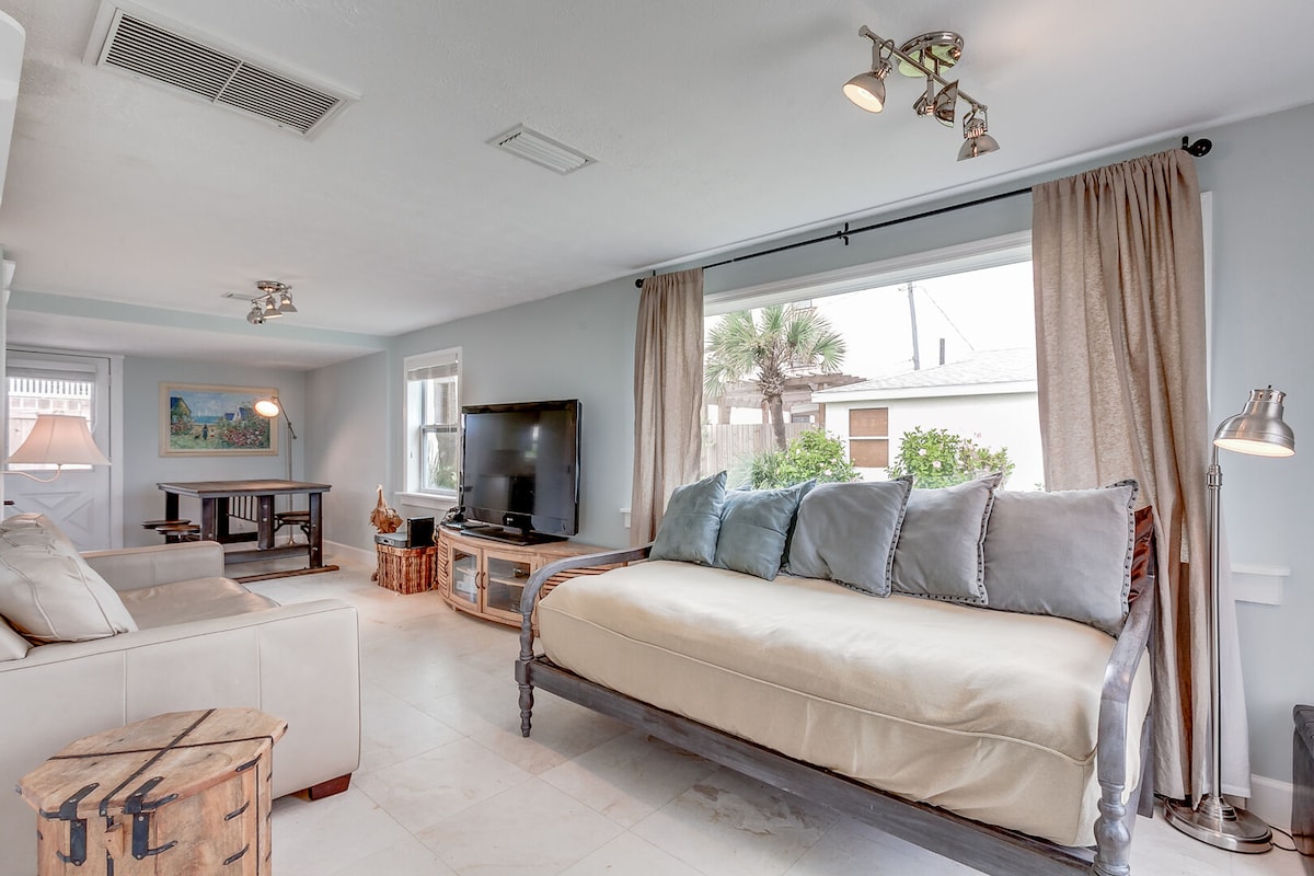 A bright living area showcases a cozy daybed adorned with plush cushions, complemented by large windows providing natural light and views of the greenery outside. A big screen TV is positioned against the wall, enhancing the inviting atmosphere.
