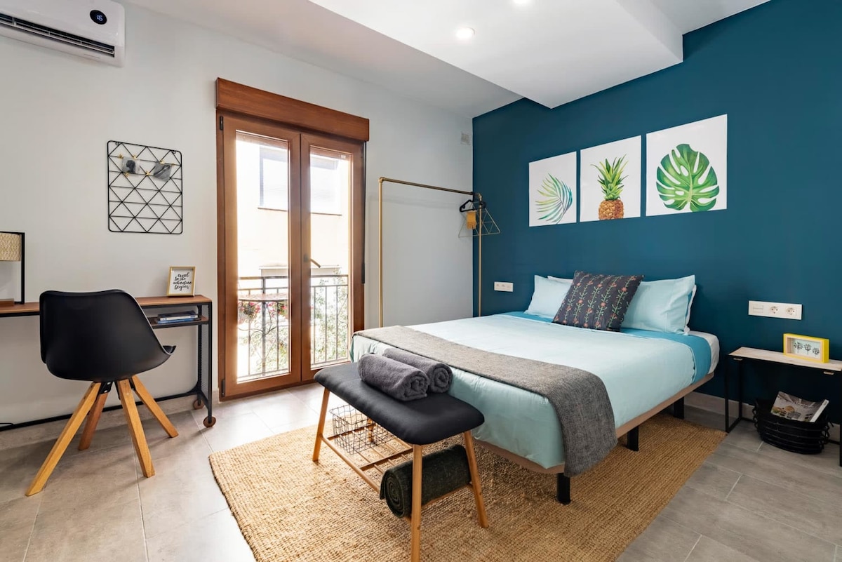 A modern studio features a spacious bed adorned with light blue linens, complemented by a decorative throw. A desk is positioned nearby, and artwork depicting plants enhances the turquoise accent wall. Natural light enters through the glass doors leading to a small balcony.