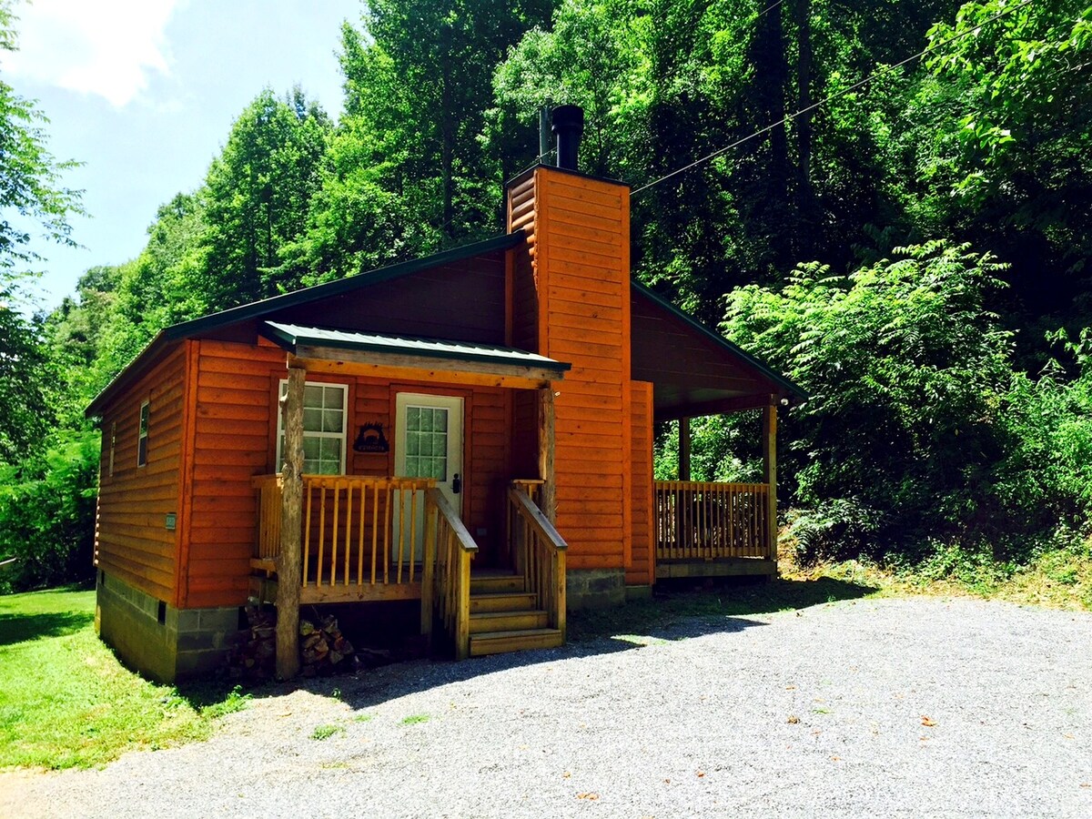 An inviting cabin with a wooden exterior is surrounded by lush greenery. The structure features a covered porch with wooden railings, a stone chimney on one side, and steps leading to the front door. A gravel driveway provides easy access.