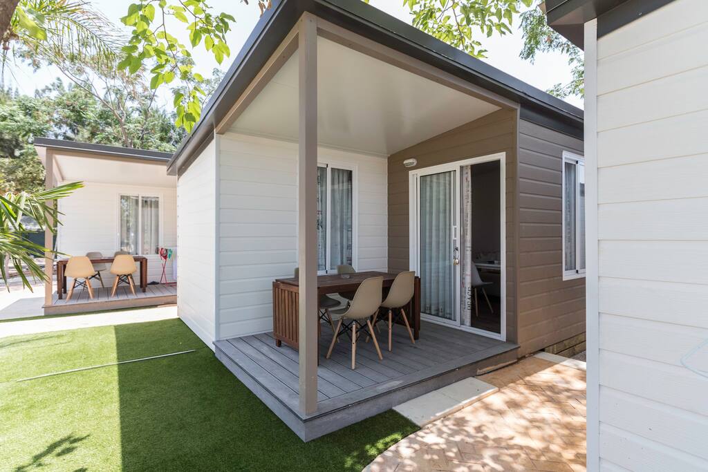 A modern bungalow exterior is shown, featuring a covered wood deck with a dining table and four chairs. Lush greenery surrounds the area, and sliding glass doors provide an entrance to the interior, enhancing the connection to the outdoor space.