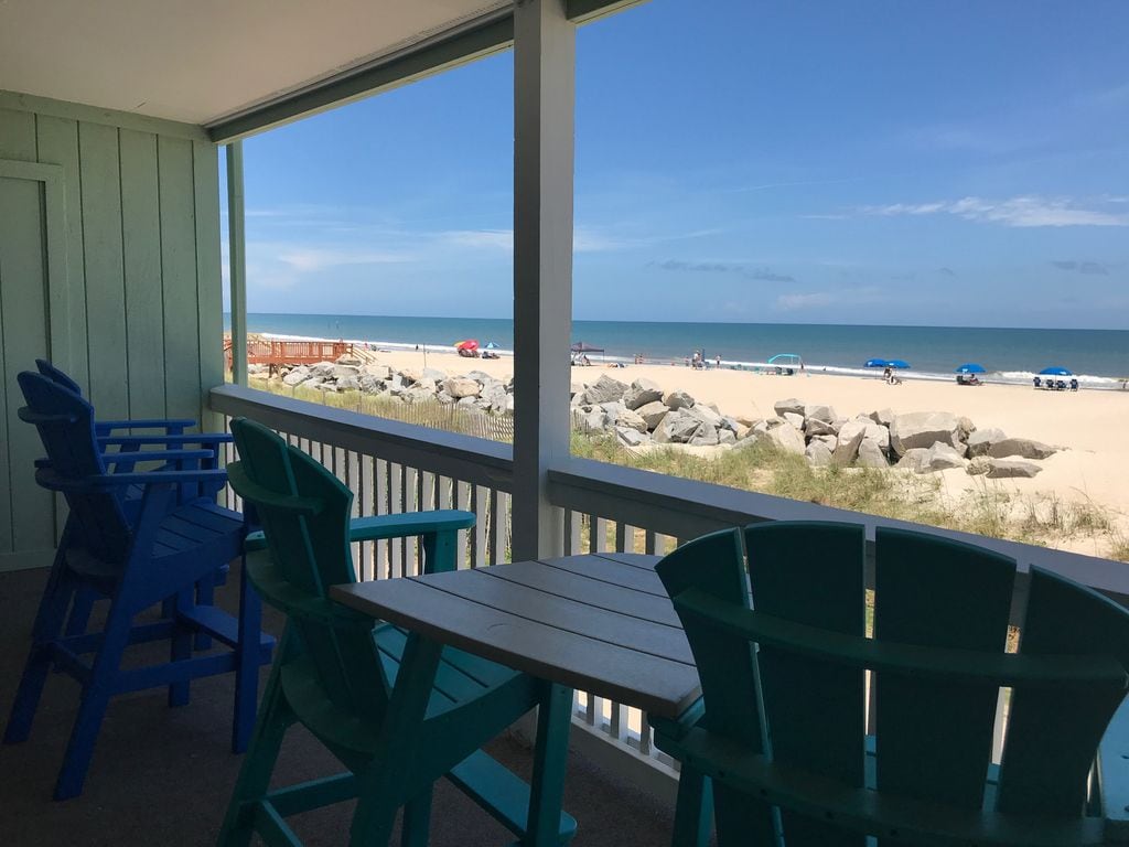 A shaded balcony offers a view of the sandy beach and ocean, with colorful adirondack chairs positioned around a small table. Rocky formations dot the beachscape, while beachgoers are visible in the distance enjoying the sunny day.