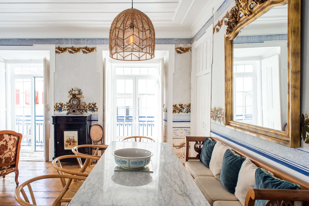 The dining area features a marble-topped table surrounded by elegantly designed Wishbone chairs. A large ornate mirror reflects natural light, while a woven pendant lamp hangs above. Decorative wall frescoes and windows framing the outdoor view contribute to the inviting ambiance.