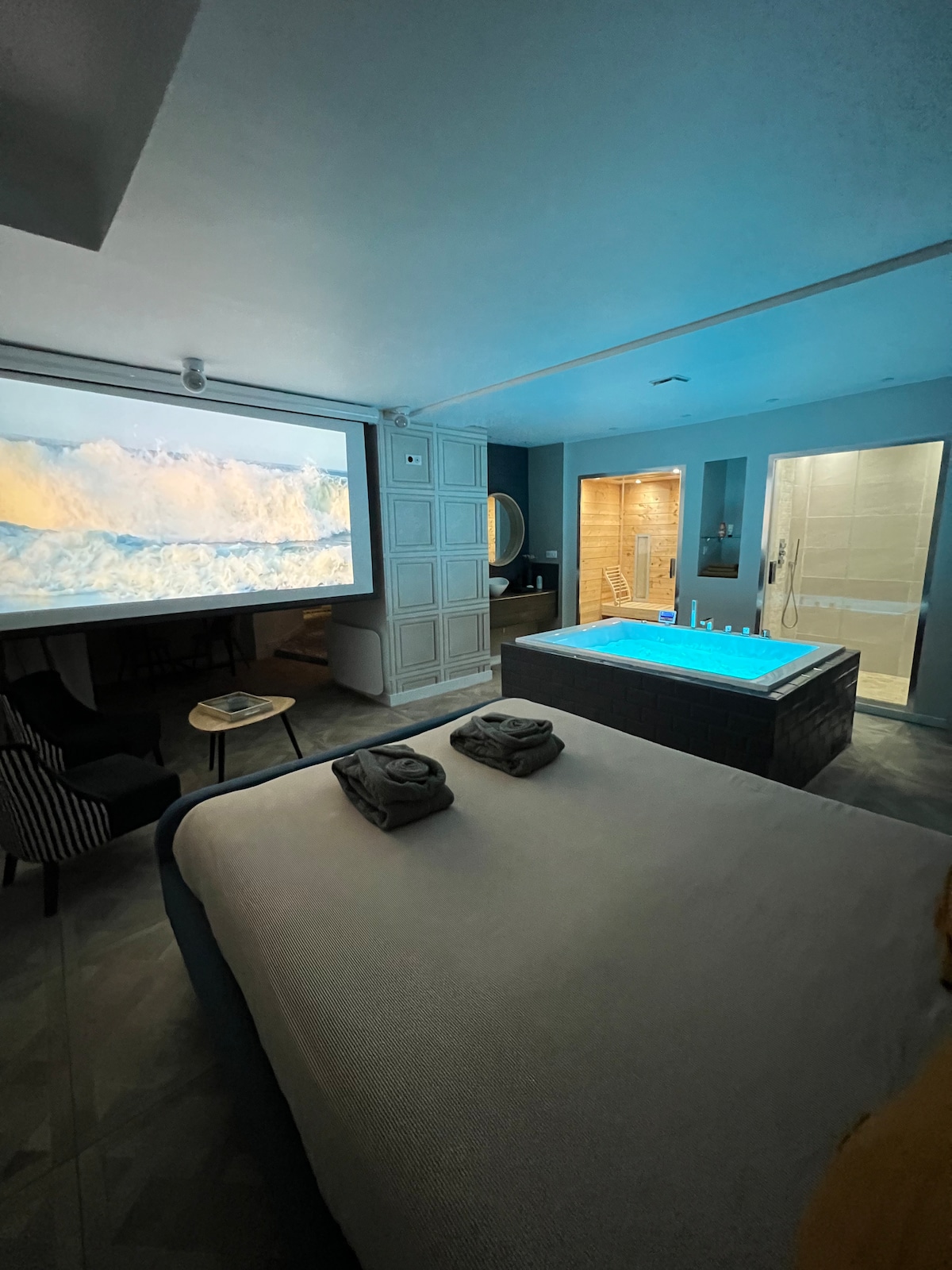 A spacious room is showcased, featuring a large bed covered with folded towels. A private spa with illuminated water is observed next to a shower area. A home cinema screen displays a coastal scene on one wall, contributing to a relaxing atmosphere.
