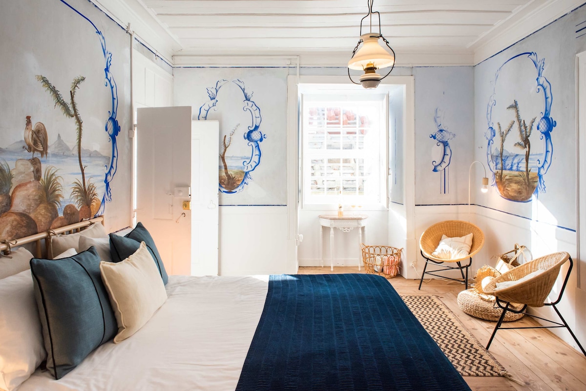 A spacious bedroom is adorned with original frescoes depicting coastal scenes. The inviting bed is dressed with soft pillows and a textured blanket, while a wicker chair and a small table are positioned near a bright window, allowing natural light to enhance the tranquil atmosphere.