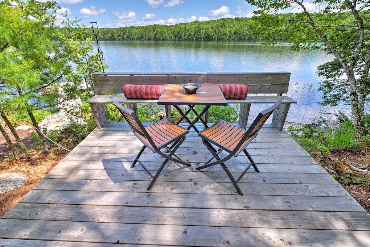 A wooden deck overlooks a serene lake, featuring a small table with two chairs. The area is surrounded by lush green trees, and a bowl sits on the table. The calm water reflects the blue sky and nearby foliage.