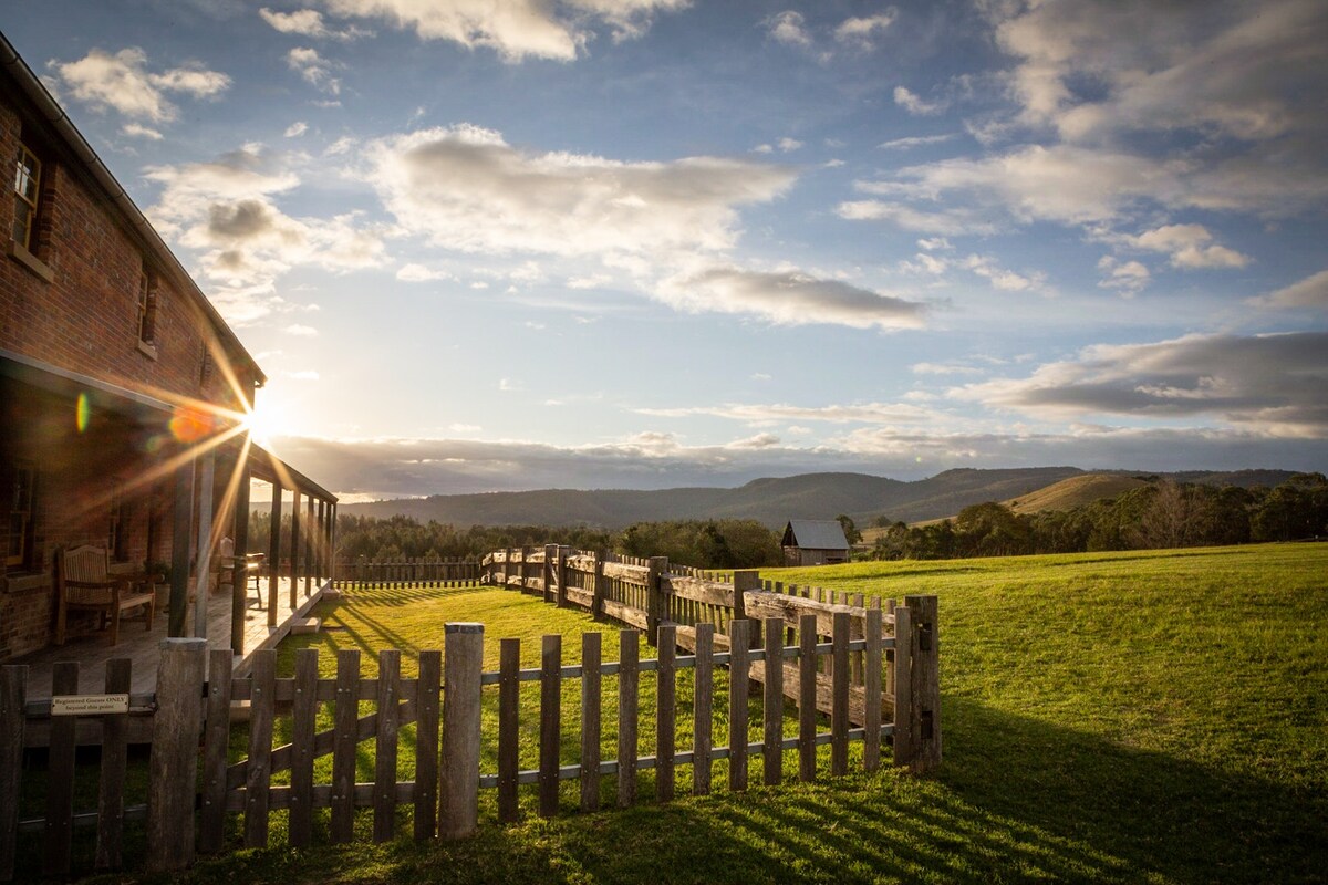 The image captures the exterior of a colonial-style building framed by a rustic wooden fence. The warm glow of the setting sun casts soft light over the expansive green fields and distant rolling hills, creating a peaceful rural landscape.