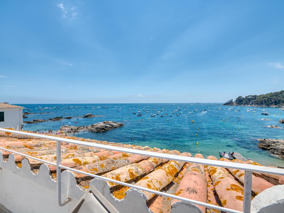 A view of the sea is displayed from the terrace, highlighting calm waters dotted with boats. The foreground features terracotta tiles with a weathered texture. The horizon is clear, framed by a bright blue sky and scattered clouds.
