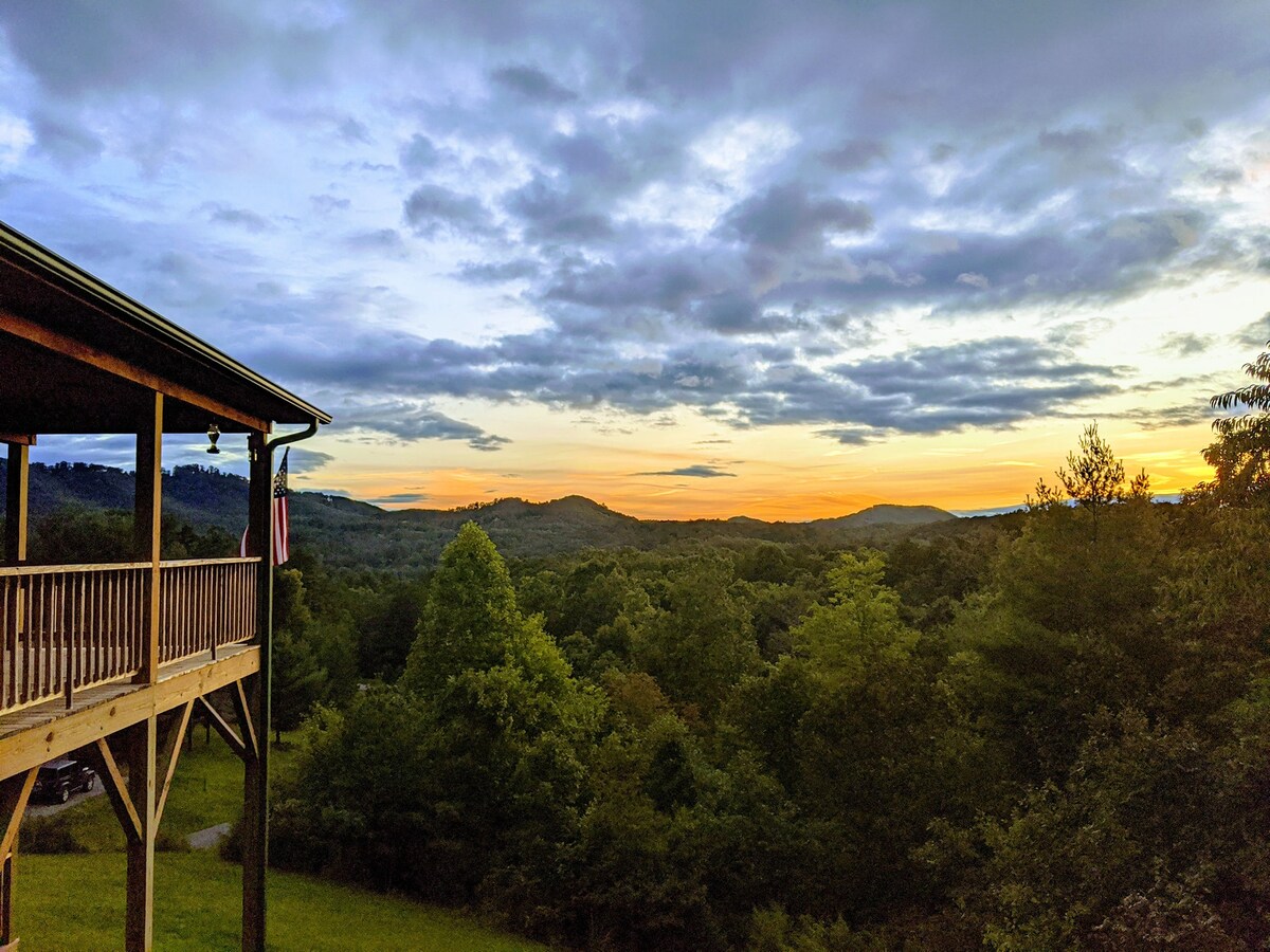 A covered porch extends from the cabin, offering panoramic views of the rolling hills and lush greenery. The sky transitions to warm hues of orange and gold at sunset, creating a serene atmosphere among the trees.