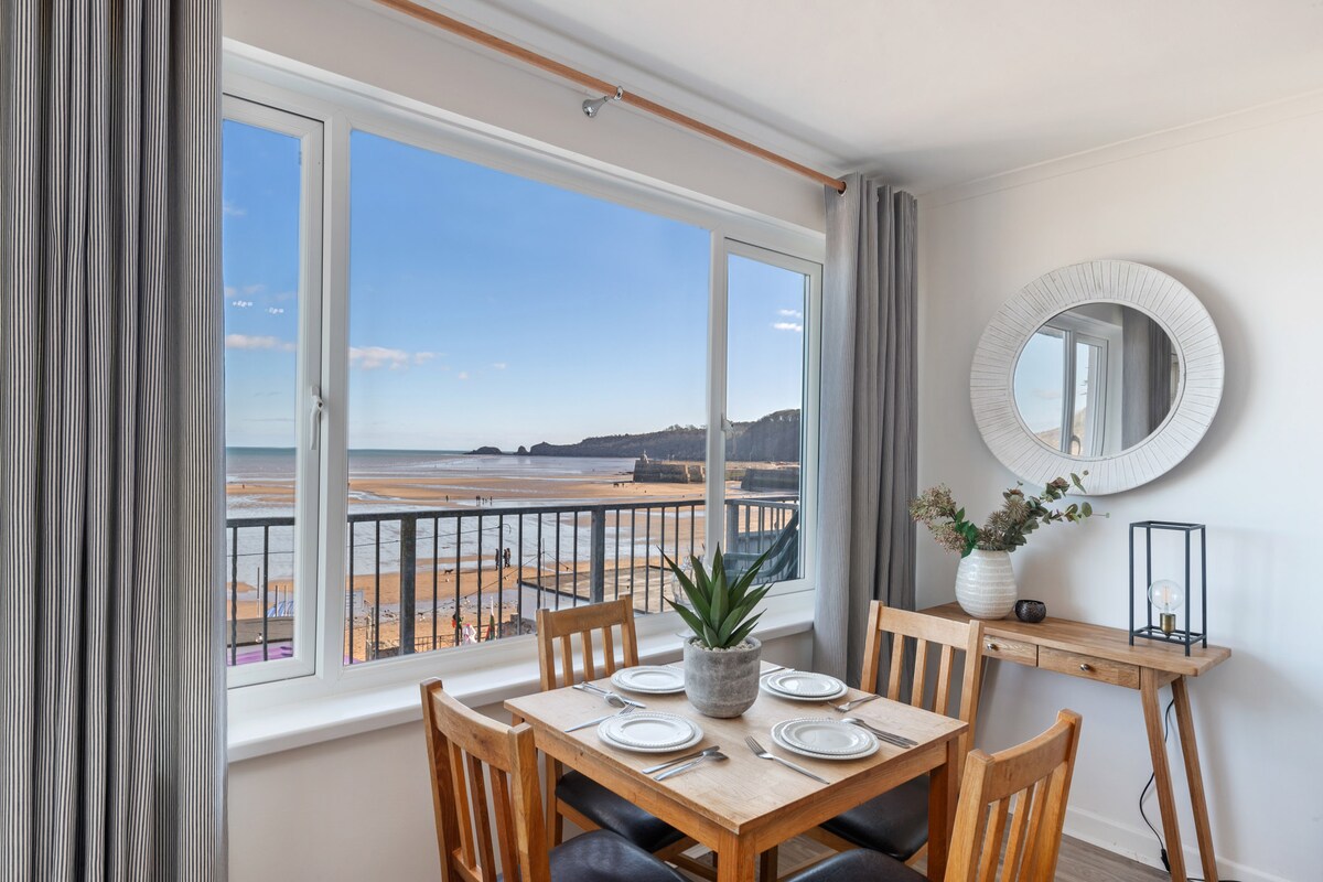 An open dining area features a wooden table set for four, complemented by a potted plant centerpiece. Large windows provide unobstructed views of the beach and coastal landscape, while soft curtains frame the scene. A round mirror and decorative lamp add subtle elegance to the space.