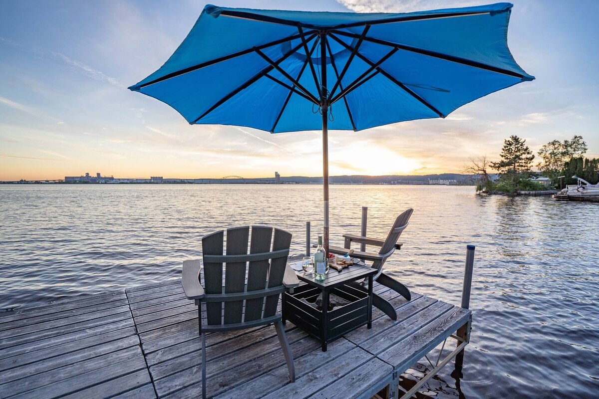 A spacious wooden dock extends over calm waters, with a bright blue umbrella providing shade for two Adirondack chairs. A small table sits between the chairs, offering a place for drinks or snacks, while a scenic sunset casts warm hues across the tranquil lake.
