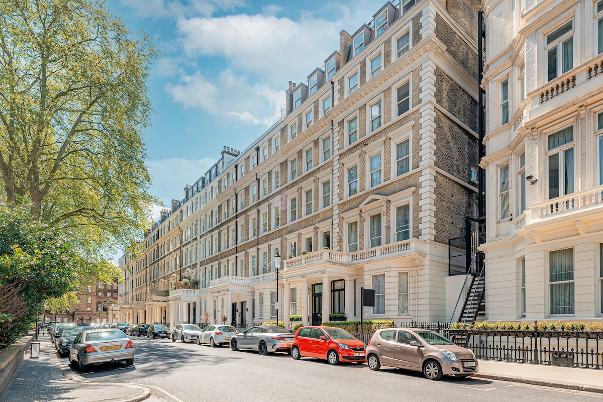 The image displays a charming street view featuring a row of elegant, multi-story buildings with classic architectural details. Trees line the street, providing greenery against the backdrop of a clear blue sky. Parked cars are visible along the curb, adding to the urban atmosphere.