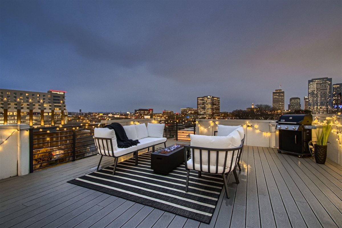An expansive rooftop with sleek outdoor furniture is complemented by soft ambient lighting. A grill is positioned next to a seating area featuring two sofas and a coffee table. The city skyline is visible in the background, providing a scenic urban backdrop.