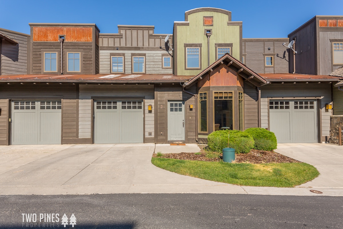 The townhome exterior features a modern design with a blend of materials, showcasing a welcoming entryway framed by large windows. The two-car garage doors are neatly arranged, and landscaped greenery adds a touch of nature to the entrance.