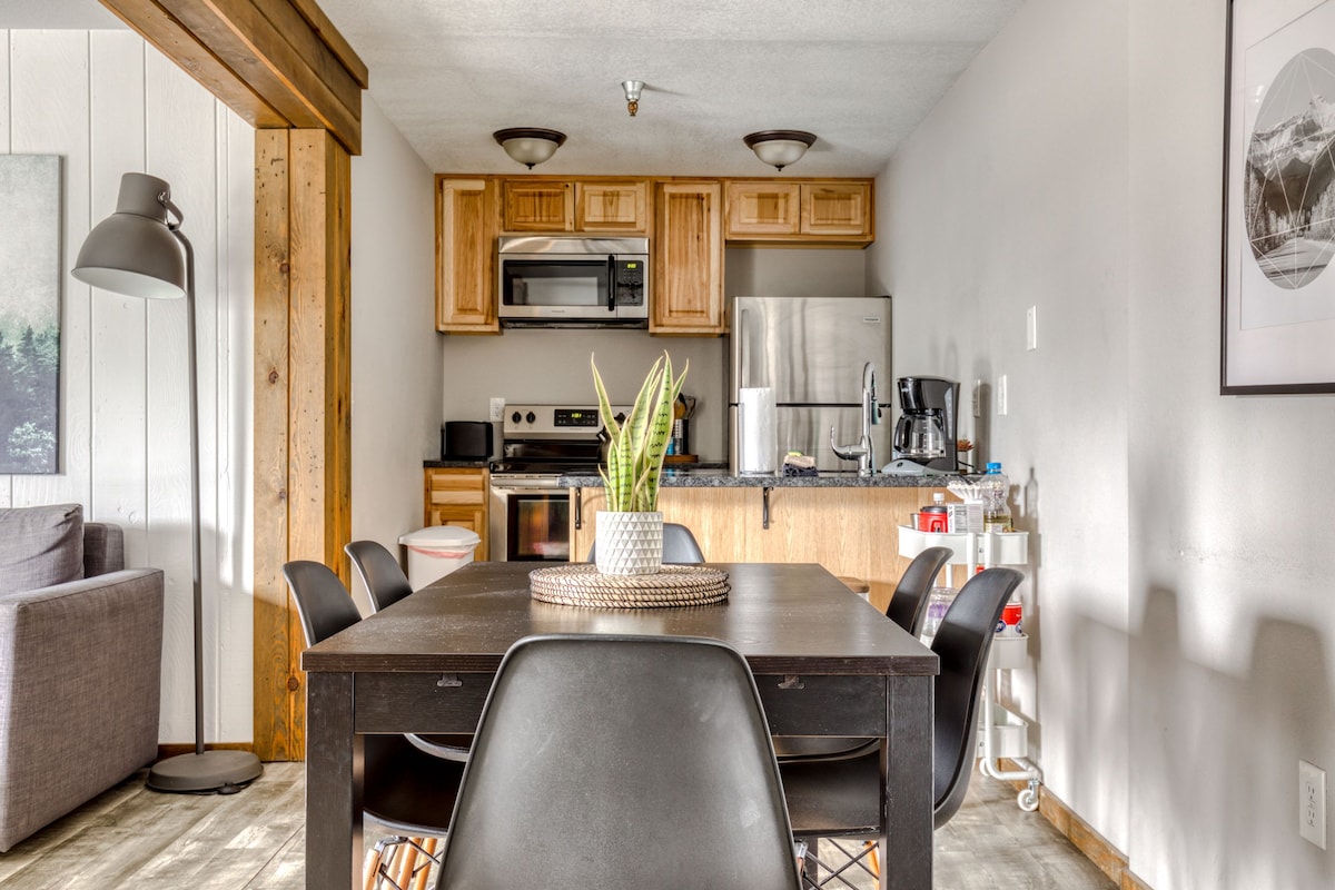 An open-concept kitchen and dining area features wooden cabinets and modern appliances. A dark dining table is surrounded by six black chairs, while a small plant adds a touch of greenery. A cozy seating area with a grey sofa is visible in the background.