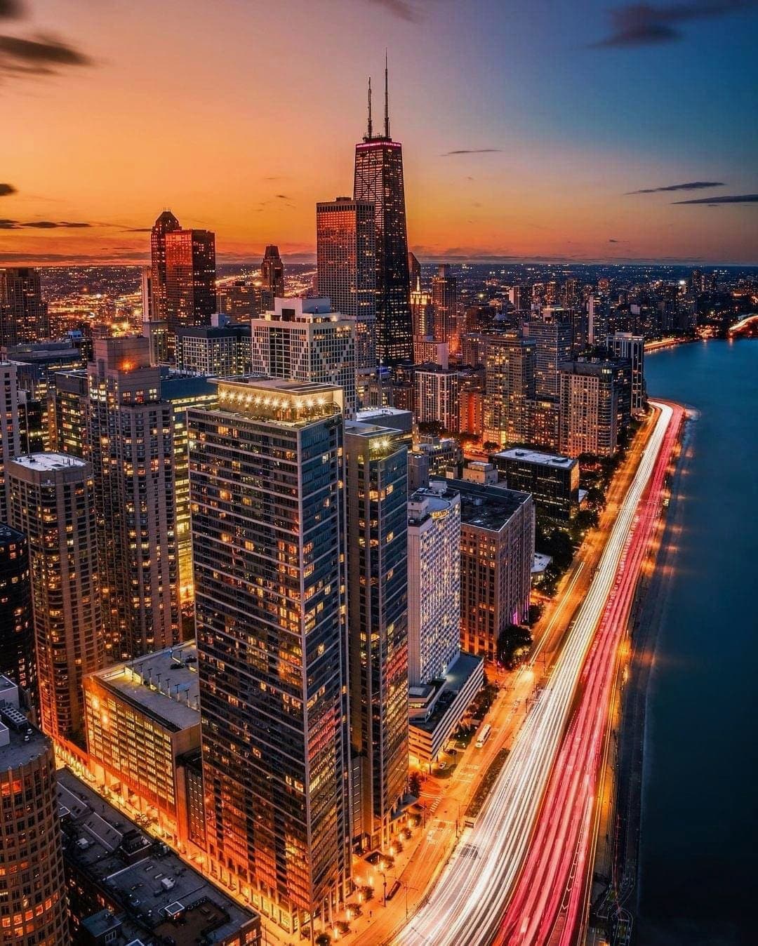 A panoramic view of a vibrant city skyline at dusk, featuring tall buildings illuminated with warm lights. The orange and purple hues of the sunset reflect on the river, while streaks of headlights from moving vehicles create a sense of motion along the waterfront.