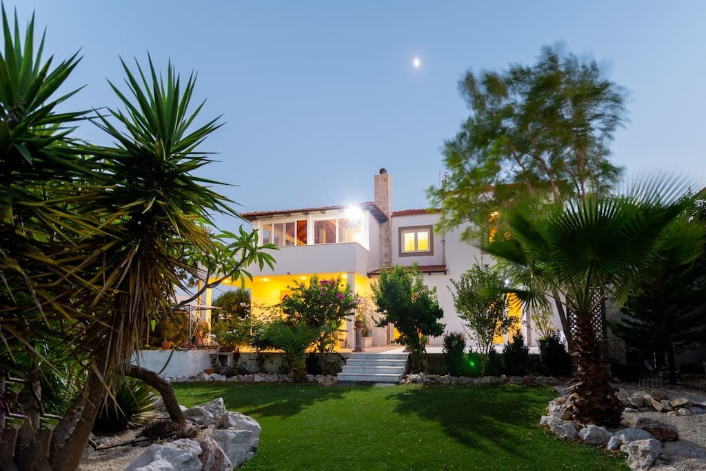 The exterior of a two-story villa is showcased amidst lush greenery, featuring palm trees and well-kept gardens. Soft lighting highlights the entrance and balconies, while the moon casts a gentle glow over the property. Stone steps lead to the welcoming entrance.