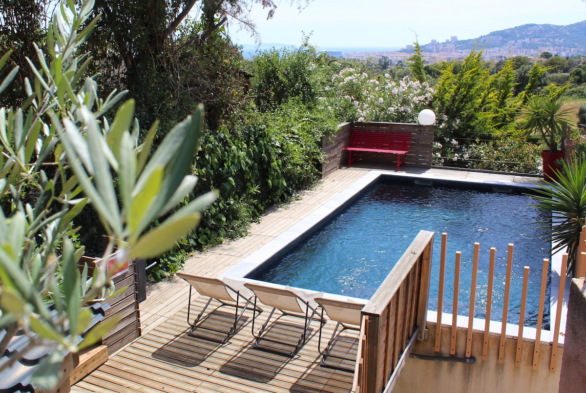 A serene outdoor pool is set within a wooden deck area, surrounded by lush greenery and flowering plants. Two sun loungers are positioned beside the pool, while distant views of Ajaccio can be seen in the background, enhancing the tranquil environment.