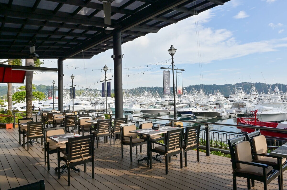 An outdoor dining area is arranged with multiple tables and chairs, overlooking a marina filled with boats. The space is sheltered by a wooden roof, with decorative lighting and a view of the serene water. The backdrop features lush greenery and distant hills.