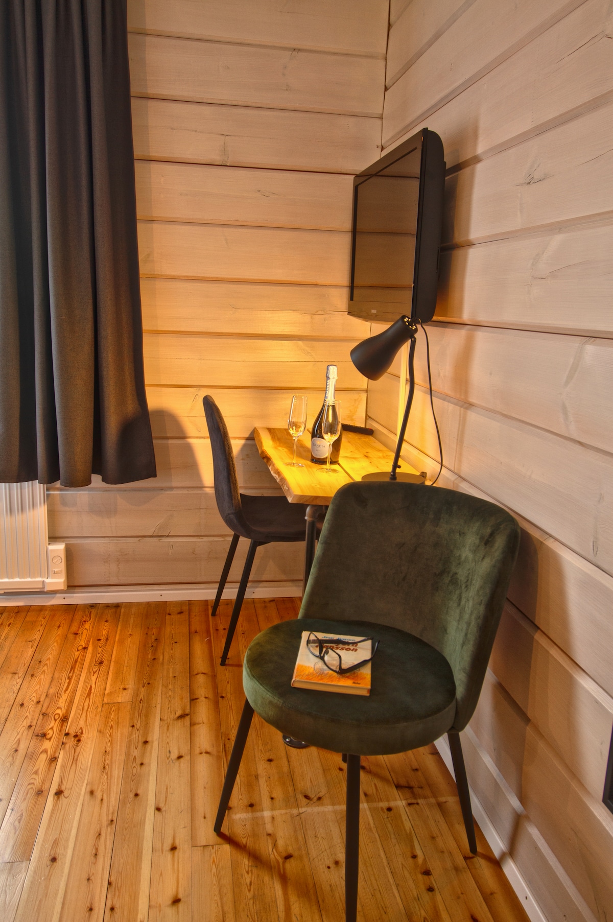 A corner of the room features a wooden desk with two glasses and a bottle of champagne, warmly illuminated by a nearby lamp. A green chair is positioned beside the desk, with a magazine resting on the surface, and a flat-screen TV mounted on the wall.