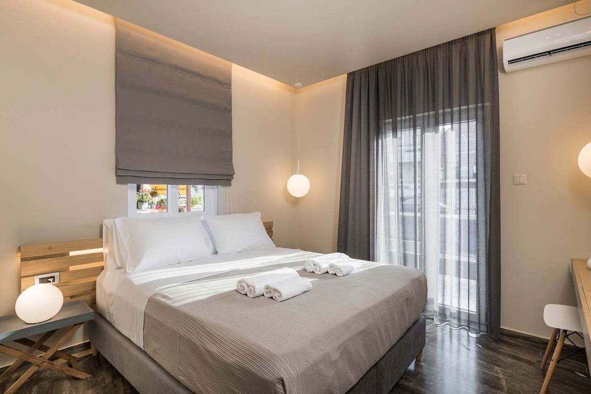 A serene bedroom features a spacious bed dressed in neutral tones, accompanied by two rolled towels. Natural light filters through sheer curtains, illuminating the contemporary design. A private balcony is accessible through glass doors, complemented by modern lighting fixtures on either side.