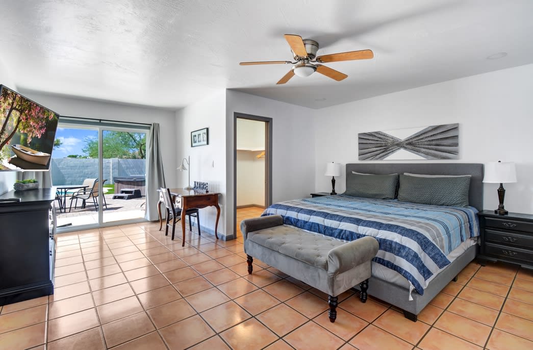 A spacious bedroom is equipped with a king-size bed featuring soft linens and a decorative throw. A ceiling fan enhances comfort, while sliding glass doors open to a private patio area. A small desk is positioned beside the bed, offering a functional workspace.