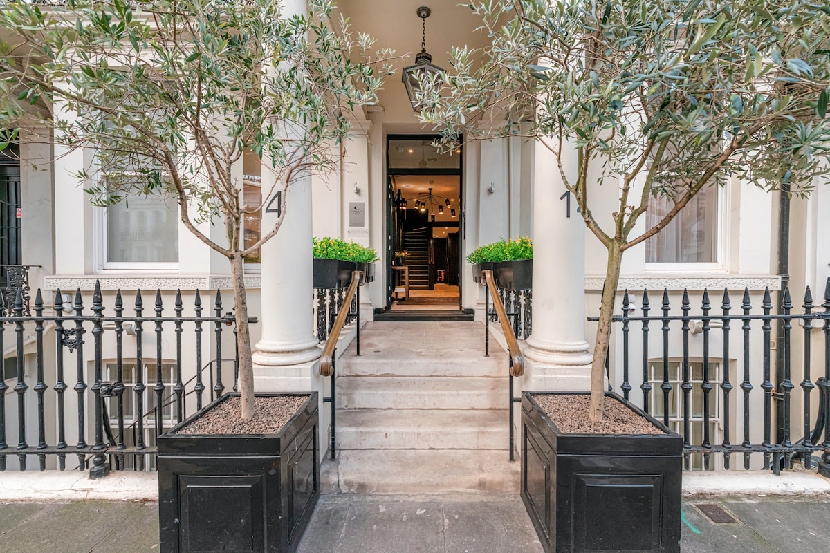 The entrance features two planters with olive trees flanking a set of wide, stone steps leading to a modern door. A wrought iron fence surrounds the property, enhancing the elegant exterior of the building.