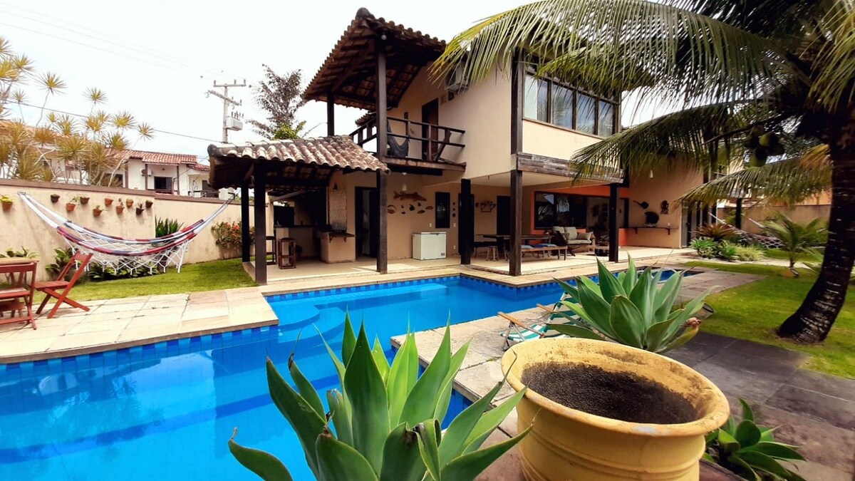 An inviting outdoor area features a sparkling swimming pool framed by tropical plants and palm trees. A spacious terrace is visible above, providing shaded seating. Nearby, a hammock and a dining area are situated under a wooden structure, enhancing the serene atmosphere of the environment.