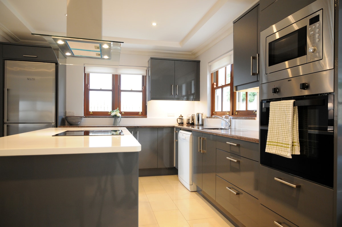 A modern kitchen features sleek grey cabinetry and a central island with a cooktop. Stainless steel appliances, including a refrigerator and microwave, are visible. Natural light enters through multiple windows, illuminating the countertop and sink area, which is equipped with essential amenities.