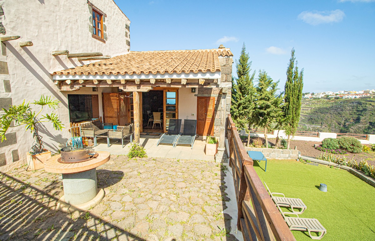 The outdoor terrace features a traditional design with a tiled roof and wooden beams. Loungers are arranged on a stone patio overlooking a landscaped area with greenery. A barbecue grill is positioned nearby, creating an inviting space for outdoor relaxation.