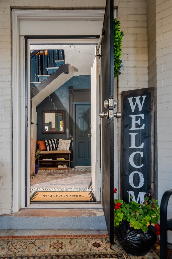 The entrance features a welcoming door with a decorative 'WELCOME' sign. A potted plant adds a touch of color beside the doorstep. Inside, a cozy area with a staircase is visible, complemented by a patterned rug that enhances the inviting atmosphere.