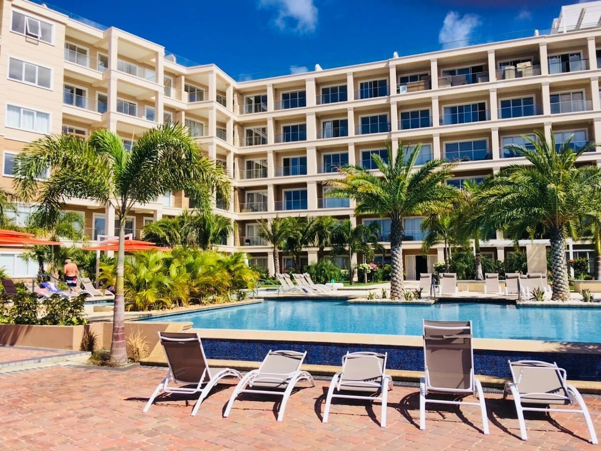 A spacious outdoor pool area is surrounded by palm trees and vibrant landscaping. Loungers are arranged along the poolside, providing a place to relax. The modern building stands in the background, featuring large windows and balconies, inviting natural light into the living spaces.