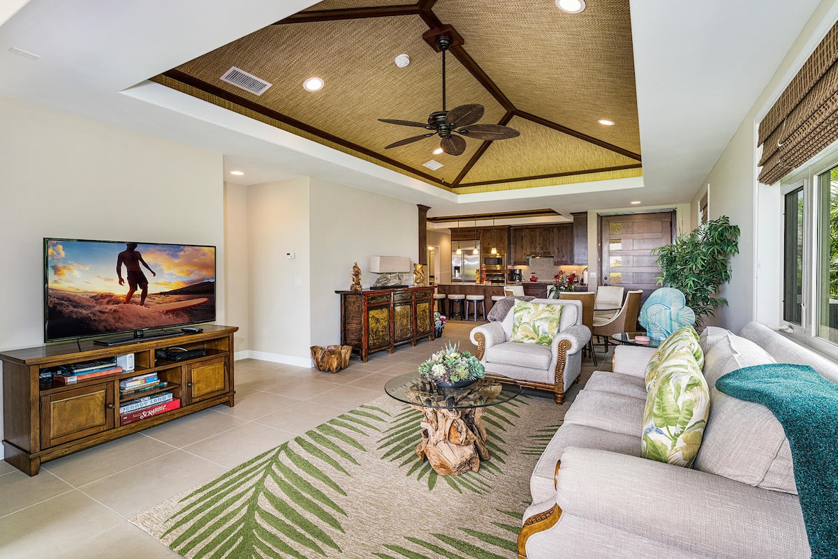 A spacious living area is highlighted by a coffered ceiling and a ceiling fan. Light-toned furniture surrounds a glass table adorned with decorative items. A television is positioned against the wall, with the kitchen and dining area visible in the background. Large windows allow natural light to enter.