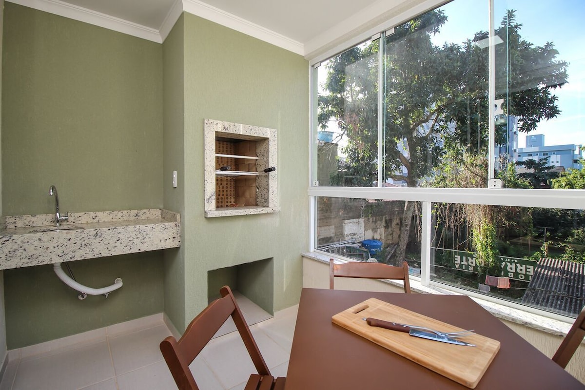 A well-lit balcony space features a barbecue grill and a stone sink, surrounded by large windows offering views of the greenery outside. A wooden table with knife and cutting board is positioned nearby, inviting outdoor dining.