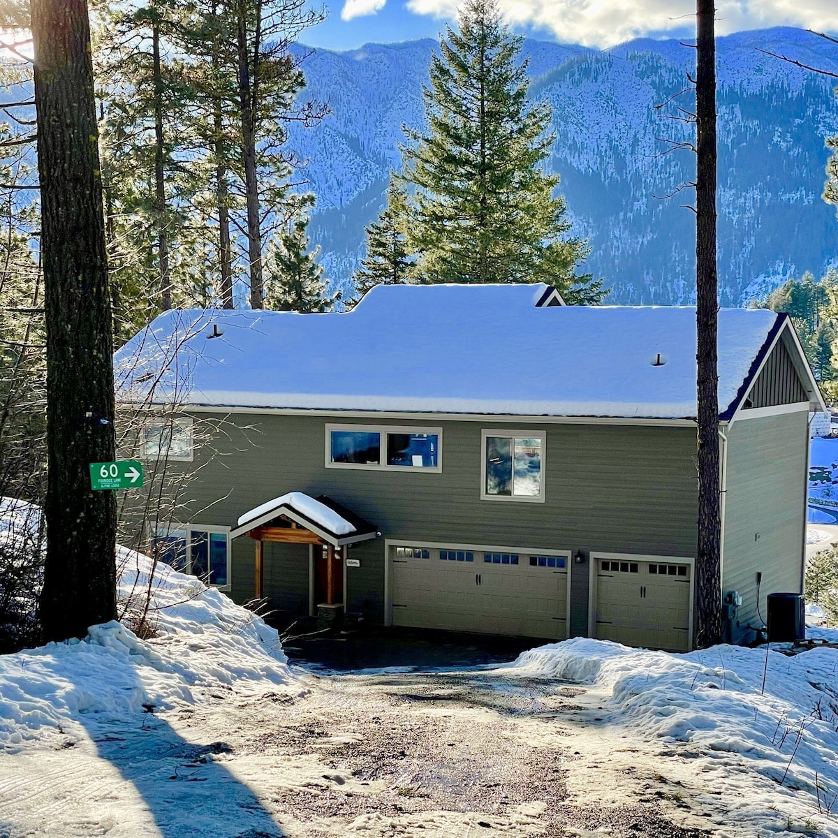 The exterior of a two-story gray home is shown, with a snow-covered roof and driveway. Large windows reflect the surrounding trees and mountains. Two garage doors can be seen, offering convenient parking. A wooden entrance is framed by fresh snow, contributing to the serene winter setting.