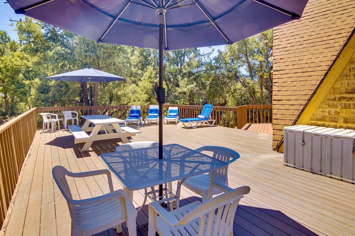 A spacious wraparound deck is featured, equipped with multiple lounge chairs and umbrellas for shade. A dining table with chairs is visible, alongside a picnic table. The deck offers views of surrounding greenery, providing an inviting outdoor space for relaxation.