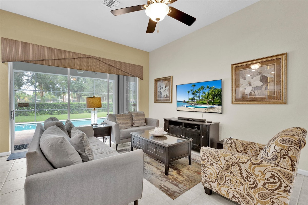 A cozy living area is furnished with a gray sectional and a patterned armchair, providing a relaxed environment. The space includes a coffee table and a media console housing a TV. Large glass doors offer views of the private pool and screened lanai.