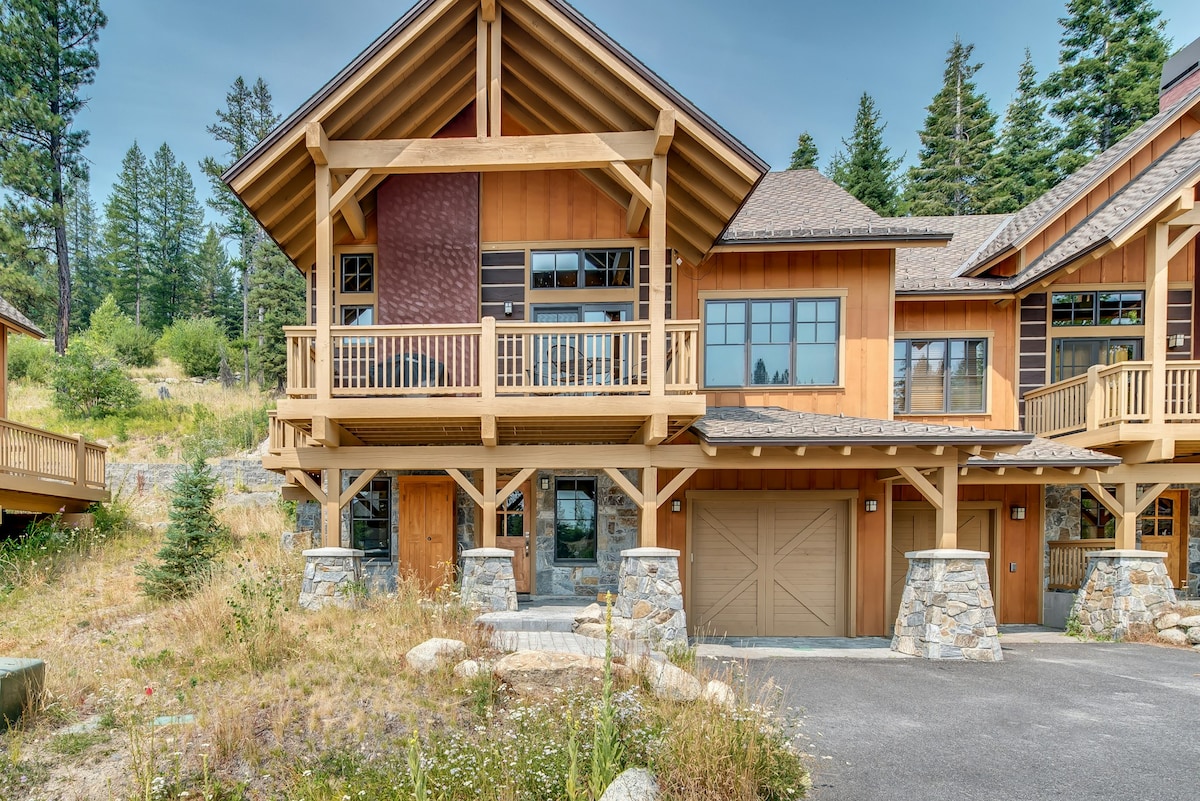 A modern cabin exterior features rich wooden siding and a large deck with railings, complemented by natural stone elements. The garage door is visible, alongside a paved driveway. Surrounding trees and natural landscaping enhance the rustic feel of the property.