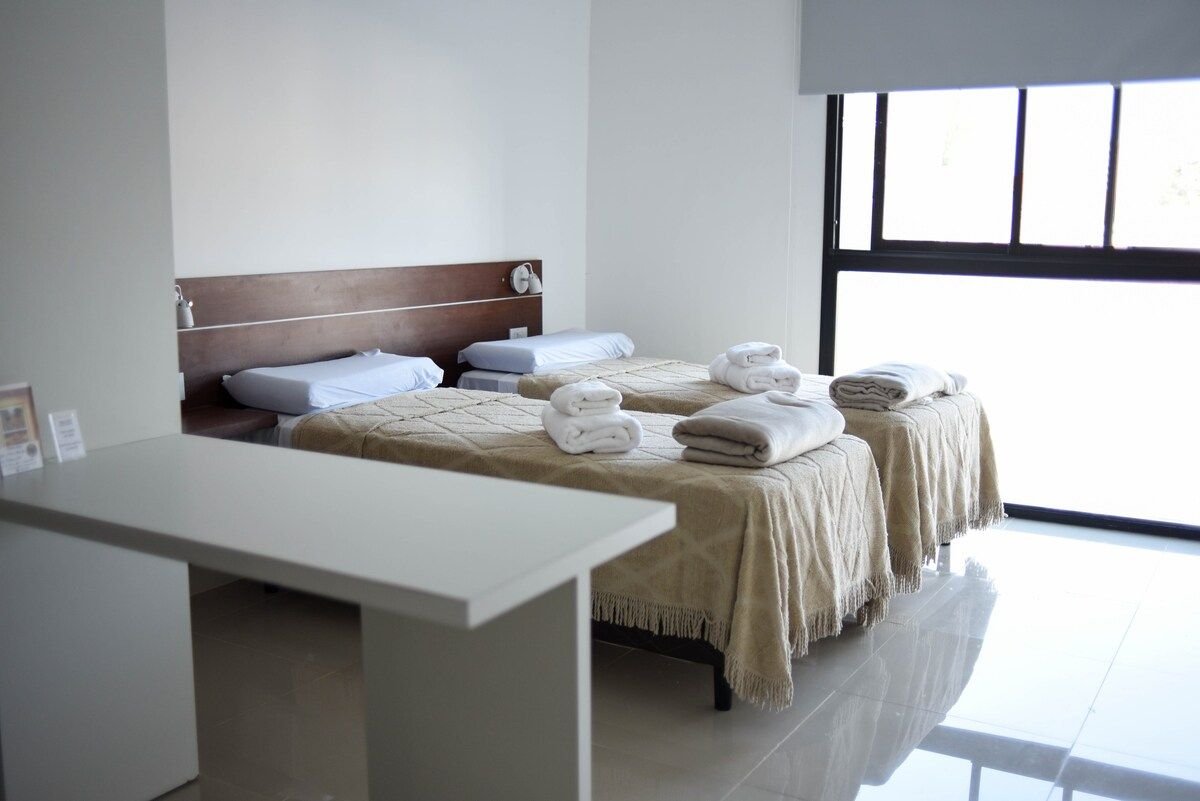 A spacious room features two beds with neutral-toned blankets, neatly arranged towels, and minimalistic furniture. A large window allows for natural light, revealing a clean, modern aesthetic.