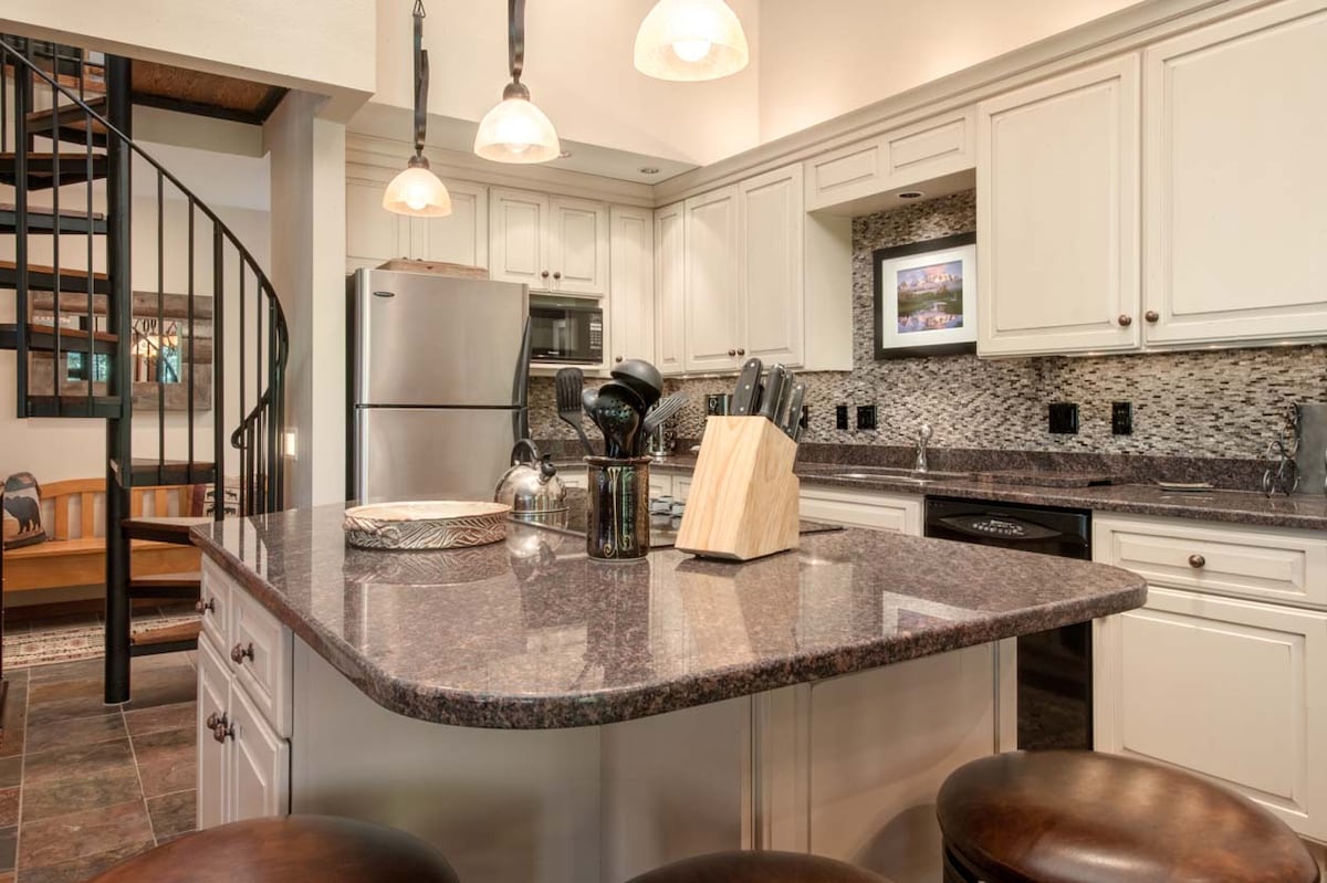 A modern kitchen features granite countertops, stainless steel appliances, and a spiral staircase visible in the background. A central island offers seating with three wooden stools. Cooking utensils are neatly arranged on the counter, complemented by warm overhead lighting.
