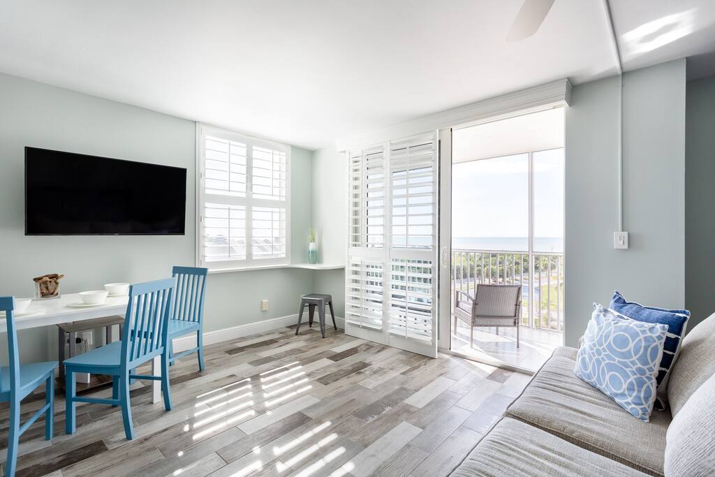 A bright living space features a large window with views of the Gulf of Mexico. A flat-screen TV is mounted on a light-colored wall. A dining table with blue chairs and a comfortable couch creates a welcoming atmosphere. Light wood flooring adds warmth to the area.