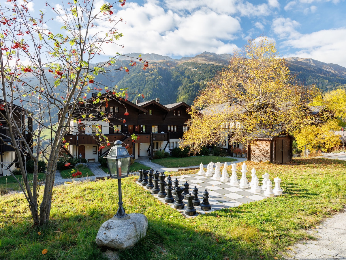 A spacious outdoor area features a large chessboard surrounded by neatly arranged oversized pieces in black and white. Nearby, trees with autumn foliage and a lamppost add a natural touch, while chalet-style buildings provide a cozy backdrop against a mountain view.