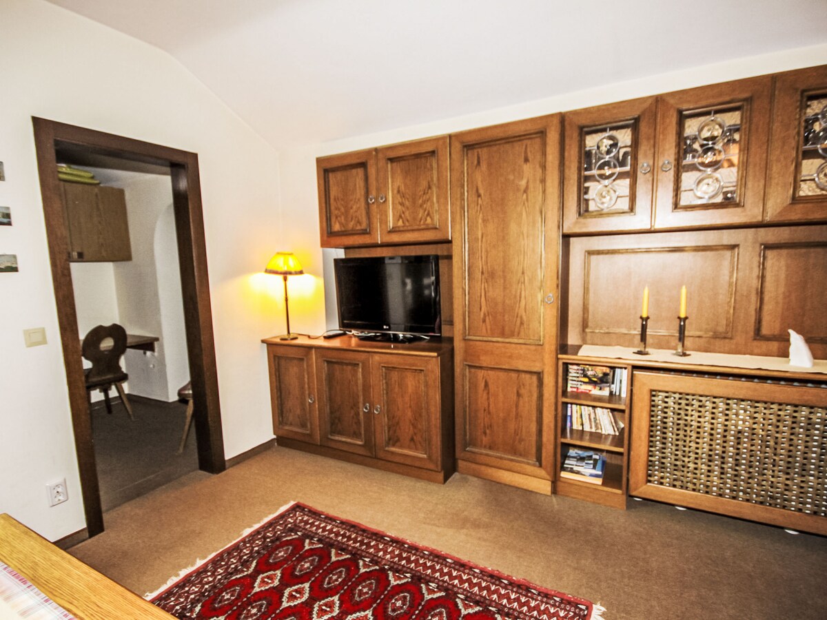 A living area is shown featuring wooden cabinetry and a flat-screen television. A warm lamp stands beside the cabinet, providing soft illumination. The carpet adds a touch of color to the neutral-toned floor, while a doorway in the background suggests access to another room.