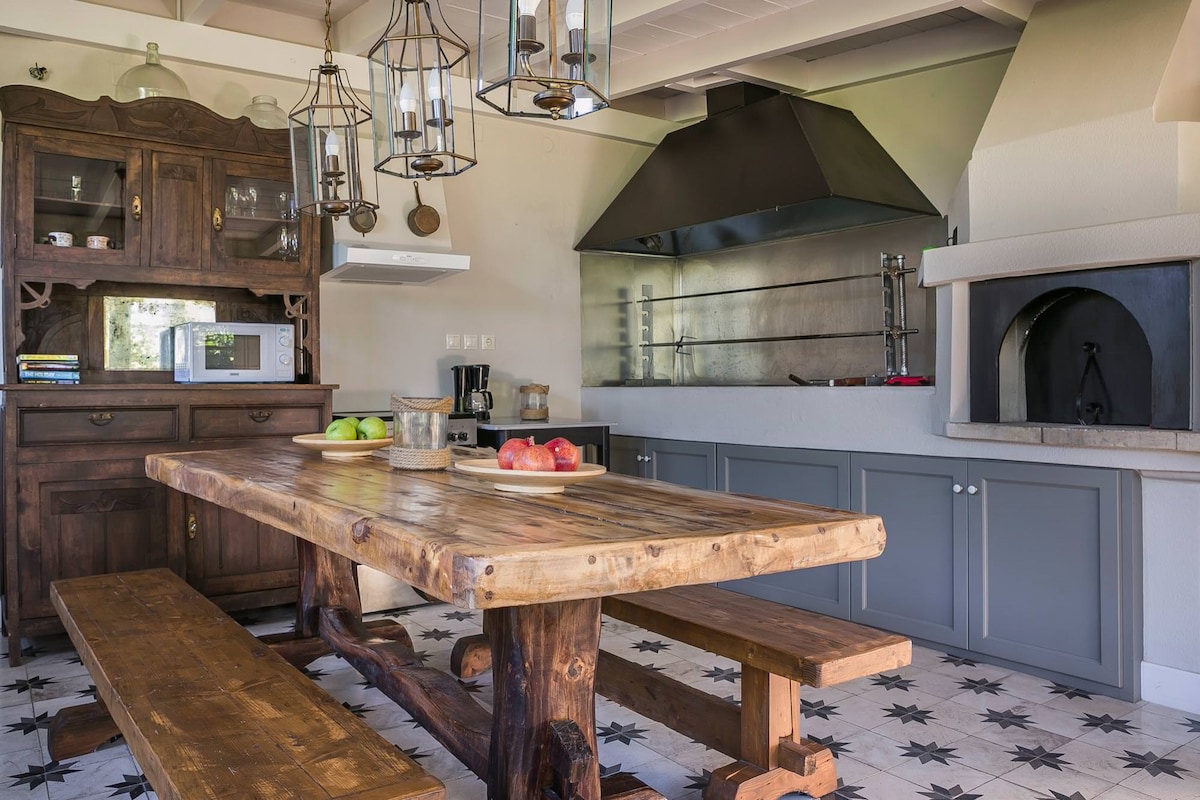 A spacious kitchen features a large wooden dining table surrounded by benches. Dark wooden cabinetry complements stainless steel appliances, including a range hood and an oven. A decorative stone fireplace adds warmth, while colorful fruit is displayed on the tabletop.