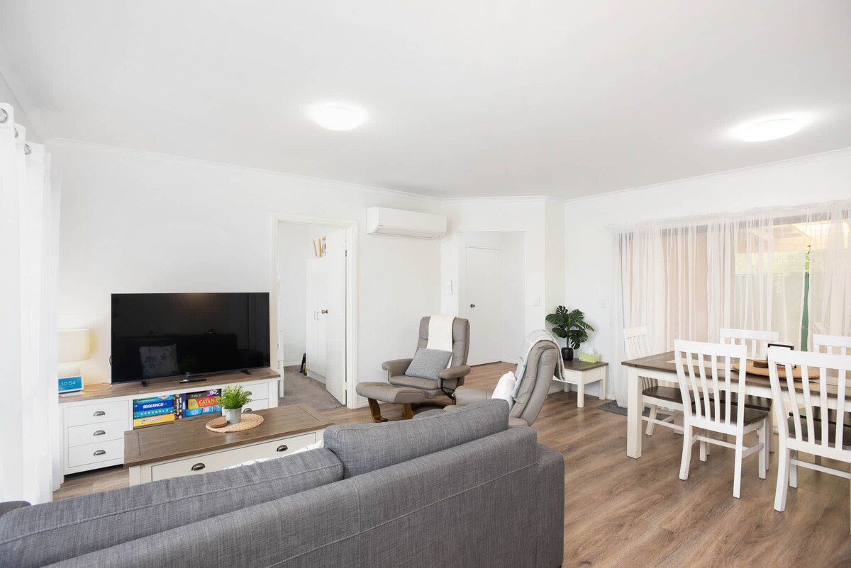 A spacious living area features a modern grey sofa and armchairs, with a large TV visible on a white entertainment unit. A wooden dining table with white chairs is positioned nearby, while a potted plant adds a touch of greenery to the space.